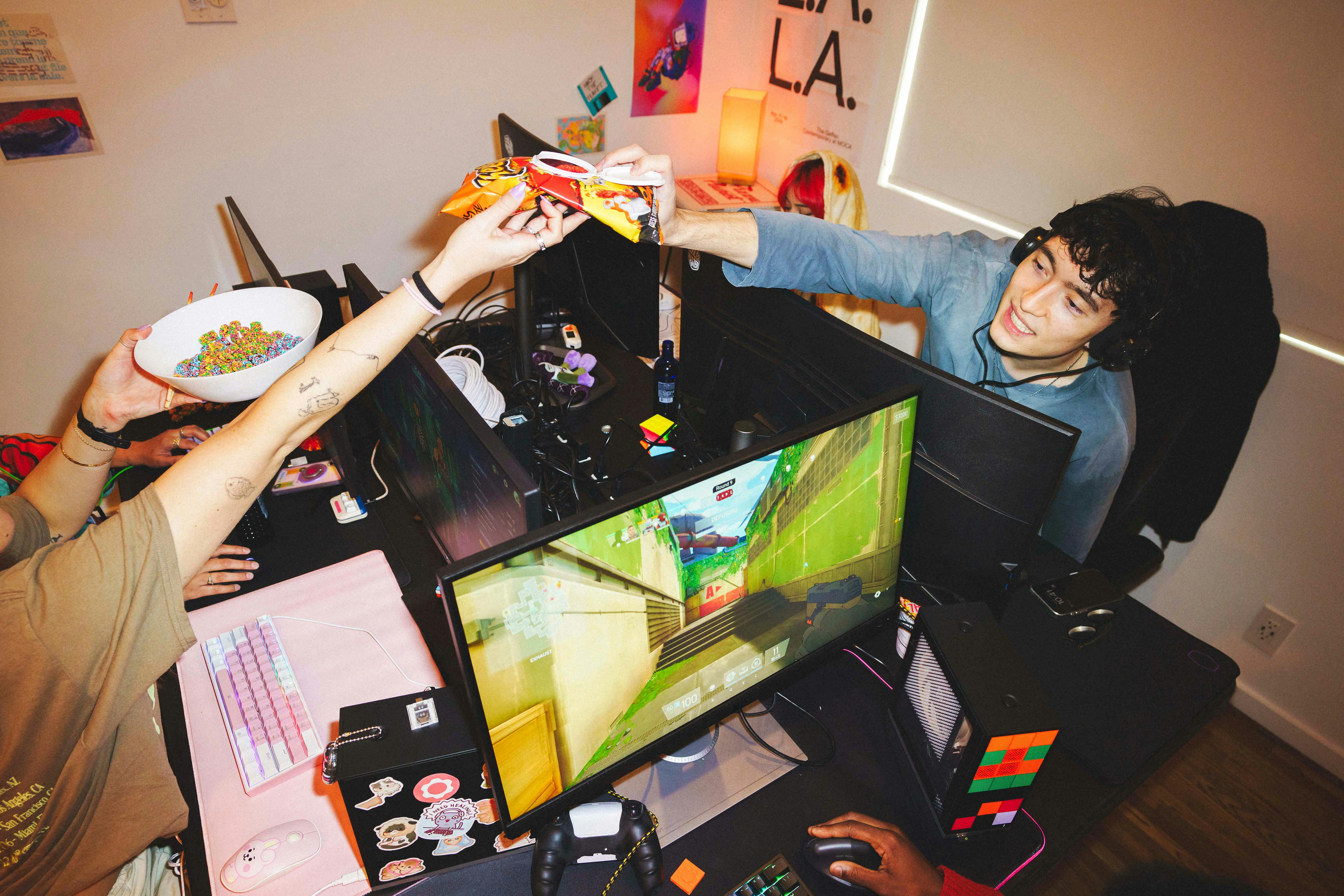 A young man wearing a headset sits in a LAN party gaming desk and reaches across to accept a bag of chips from a friend. Multiple computer monitors, a mechanical keyboard, a white bowl filled with colorful cereal, and a few compact Framework Desktops are visible.