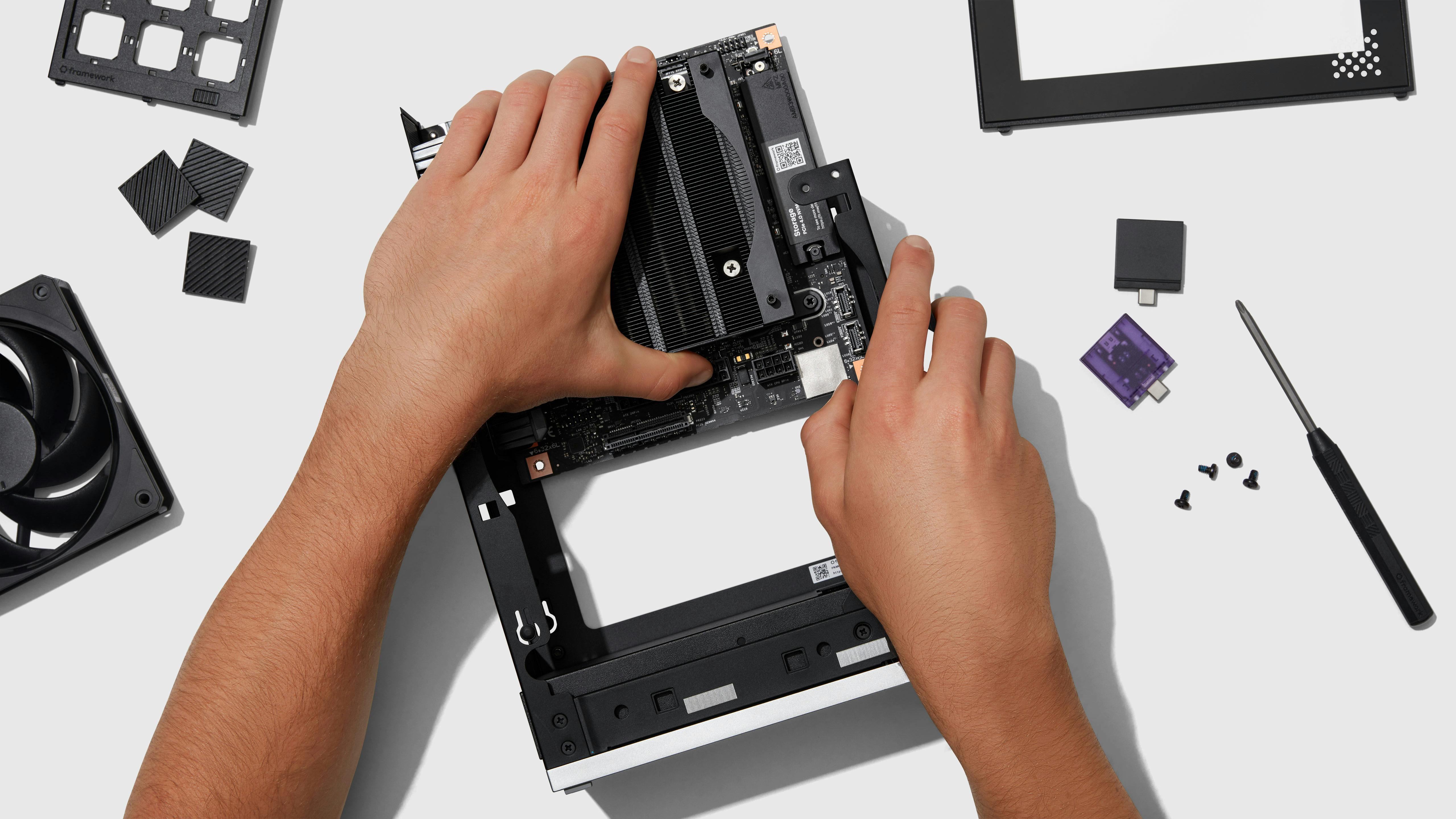A top-down view of a person assembling the modular Framework desktop chassis They are installing a mainboardcontaining a large CPU heatsink into the black frame. Scattered around the workspace are various computer components, including a cooling fan, modular expansion cards, loose screws, a small screwdriver, and frame panels.