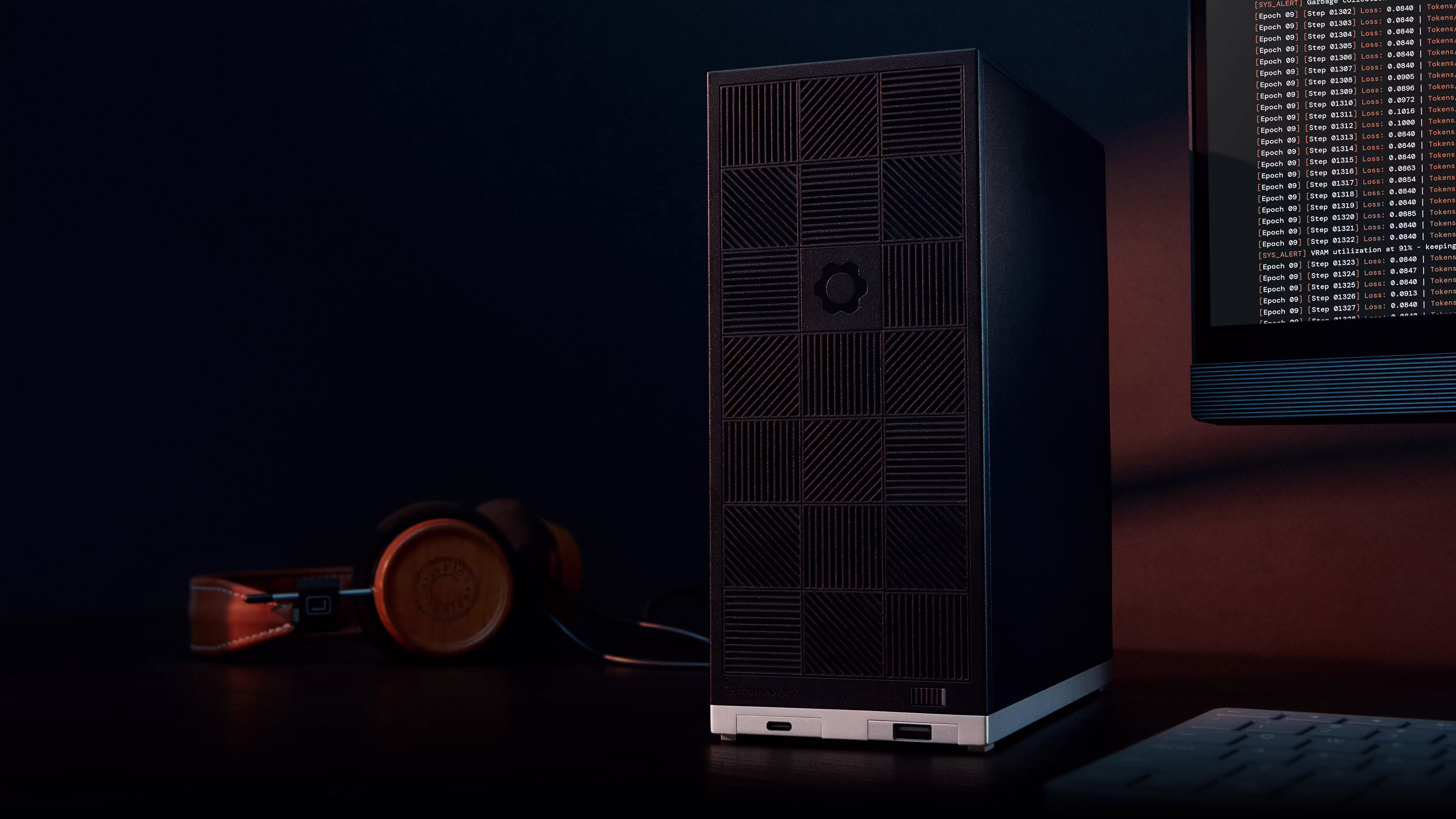 A sleek Framework Desktop stands on a dark desk under moody, low-key lighting. In the blurred background, a pair of wooden-eared headphones rests on the left, and a monitor displaying lines of code is visible on the far right.