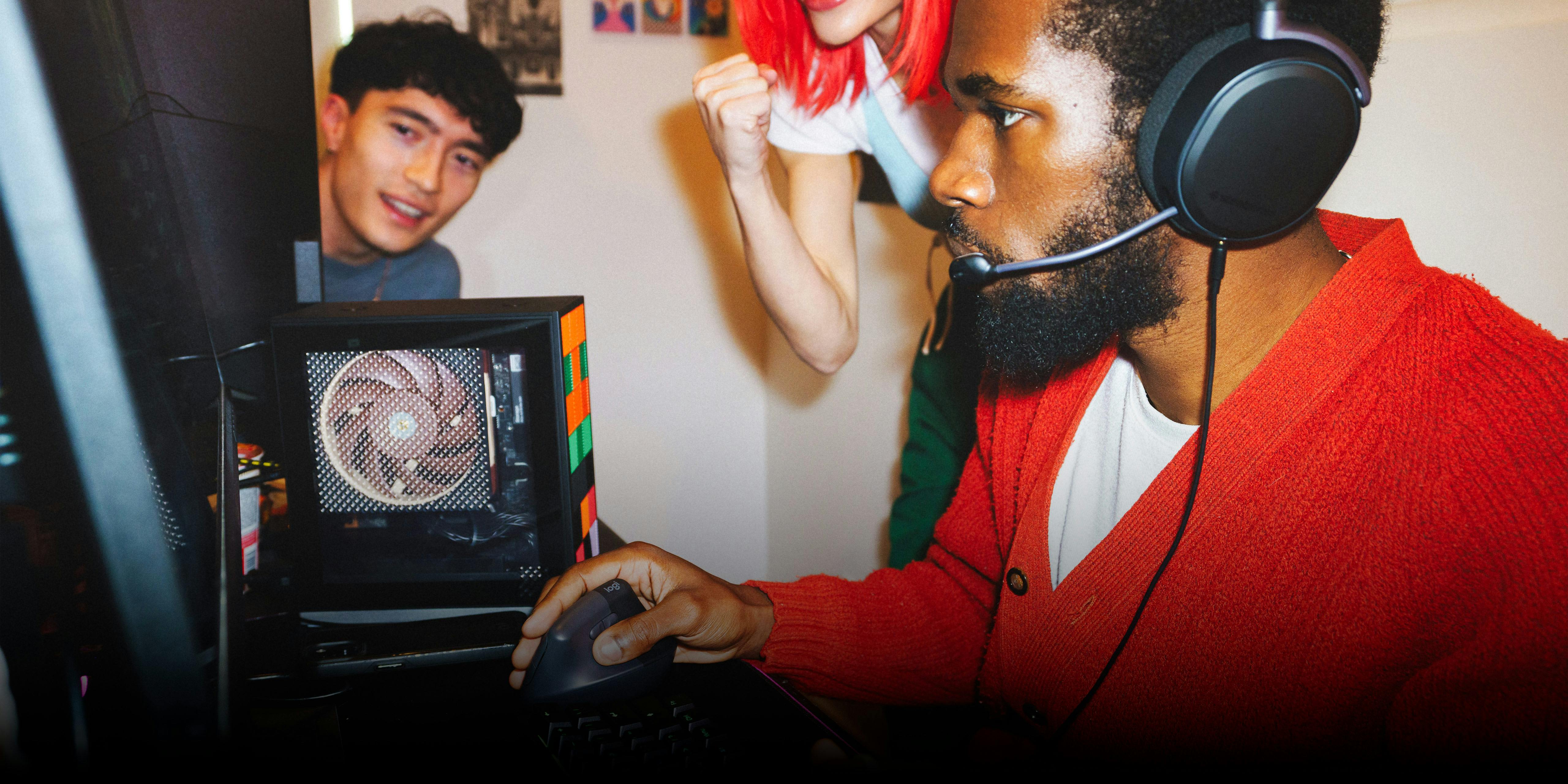 A man wearing a headset is gaming in a LAN party while two friends watch from behind. A compact Framework Desktop sits on the desk.