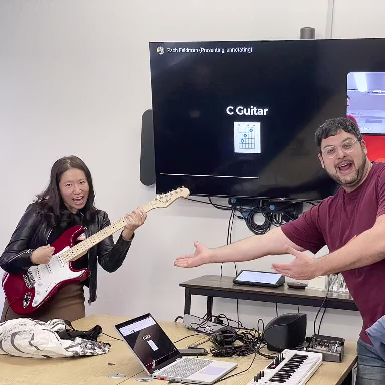 An employee holding a red electric guitar poses like a rockstar while a colleague gestures enthusiastically towards her.