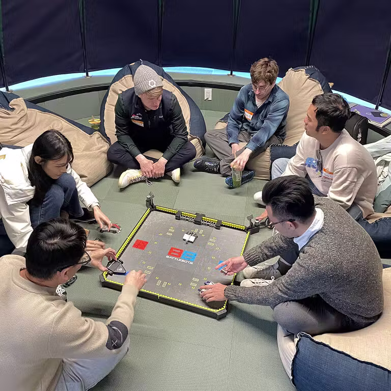 A group of Framework employees sitting in a circle, playing a tabletop game with miniature robots in a BattleBots arena.