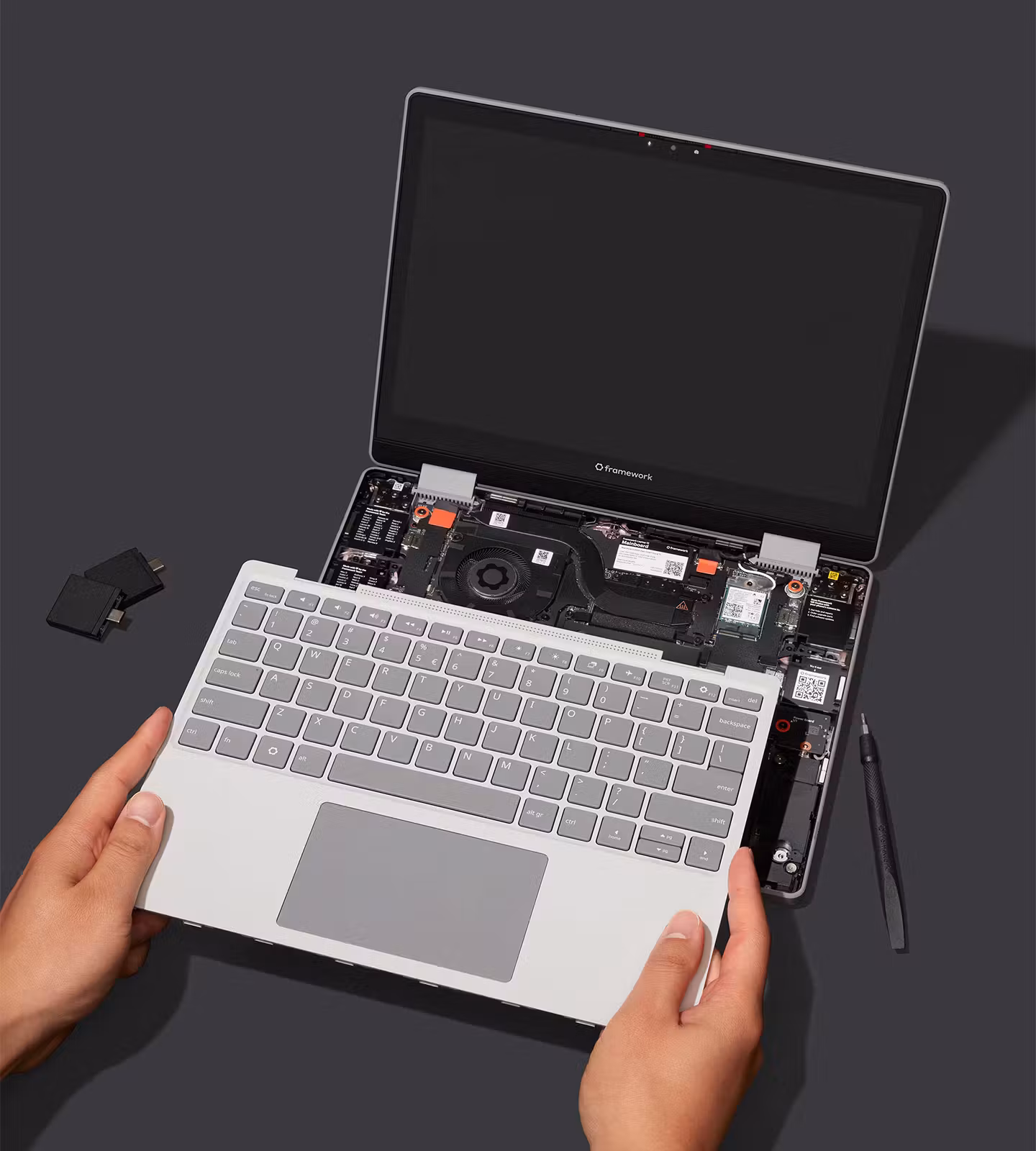 Black Framework Laptop 12 with the input cover lifted up by two hands, revealing the internal components including the fan, motherboard, and storage, with small expansion card modules beside it on a dark background.