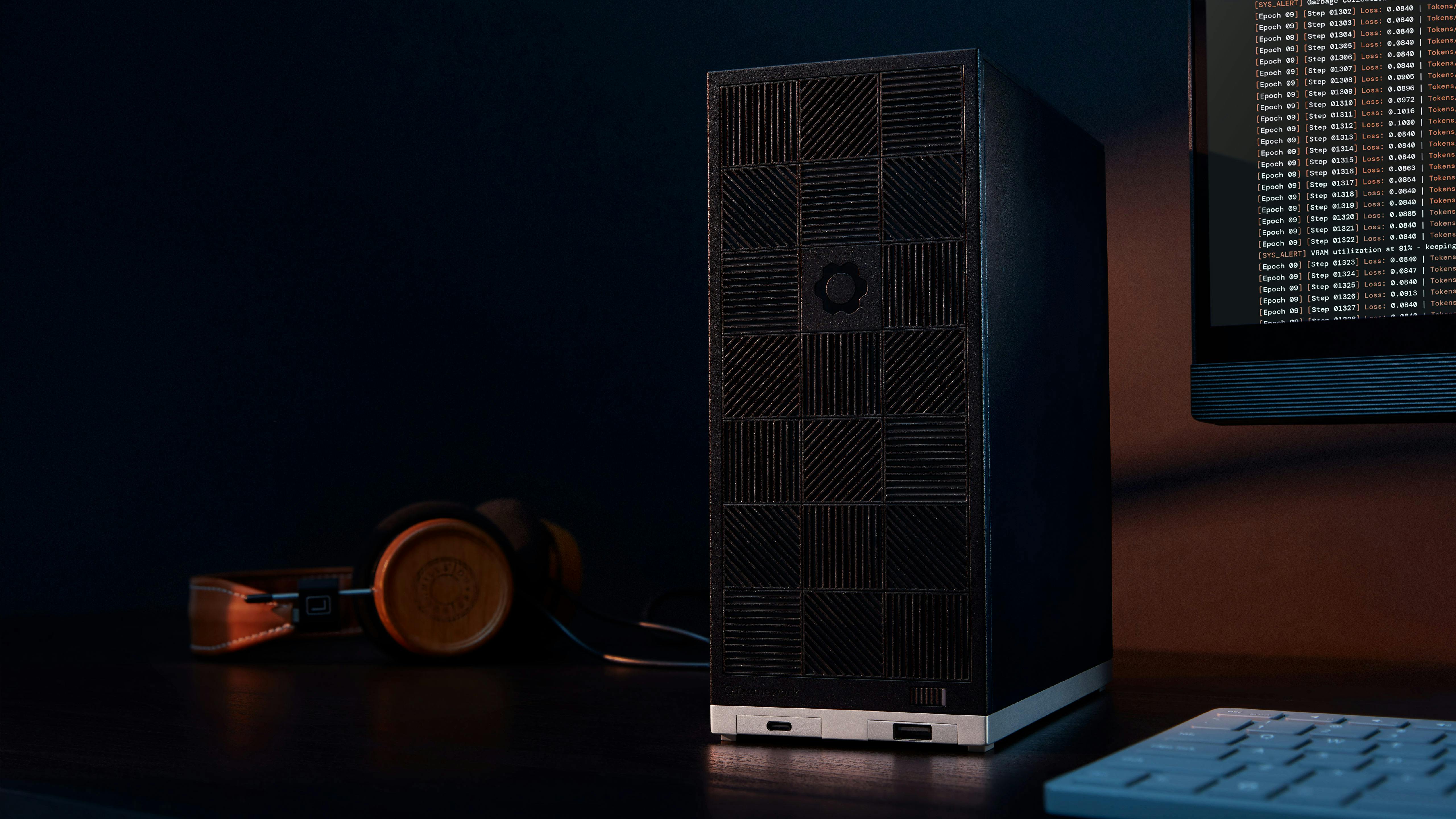 A sleek Framework Desktop stands on a dark desk under moody, low-key lighting. In the blurred background, a pair of wooden-eared headphones rests on the left, and a monitor displaying lines of code is visible on the far right.