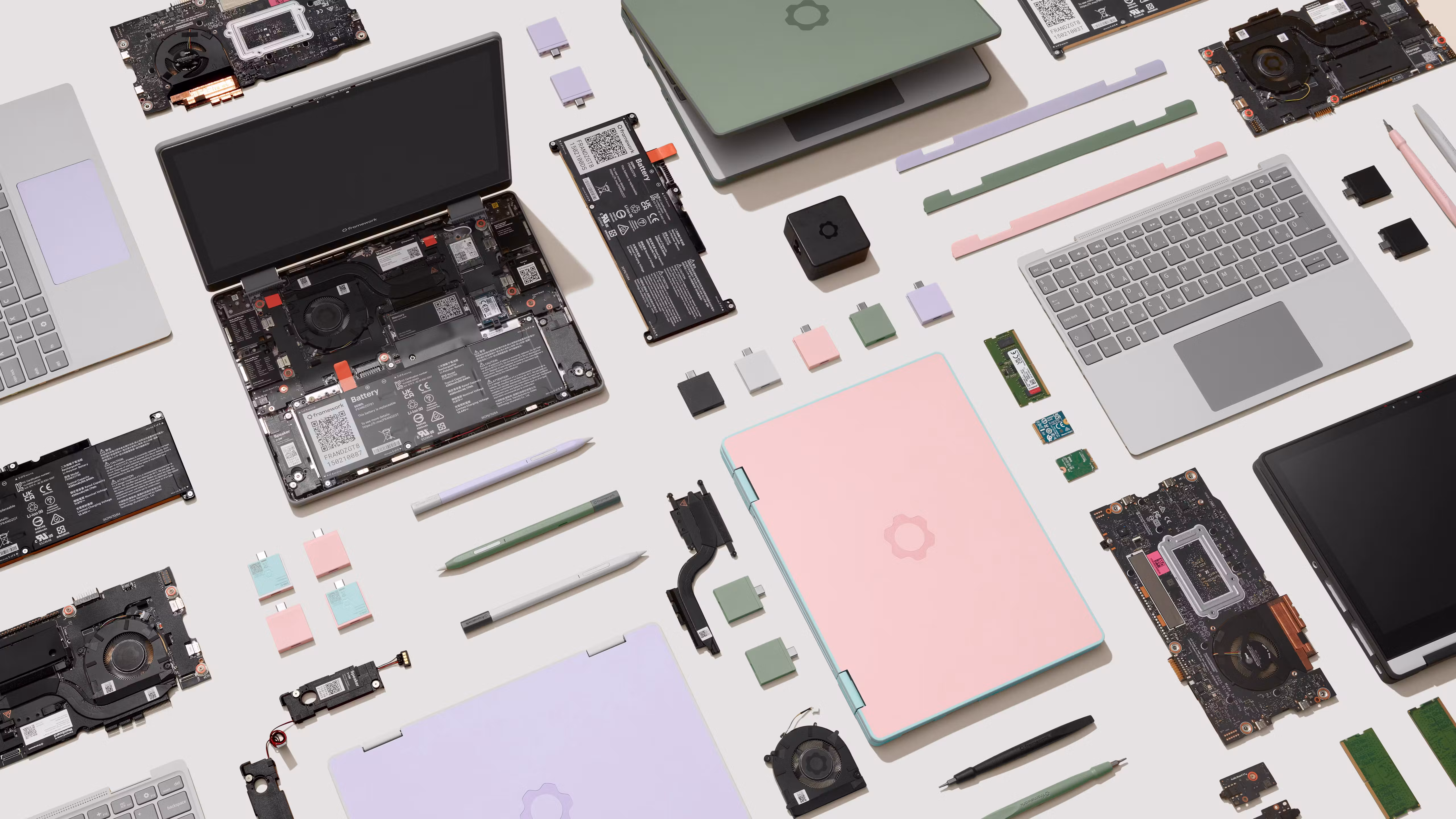 Flat-lay of many disassembled Framework Laptop 12 components spread across a table, including mainboards, batteries, keyboards, trackpads, styluses, fans, expansion cards, and laptop covers in Sage, Bubblegum, and Lavender colorways.